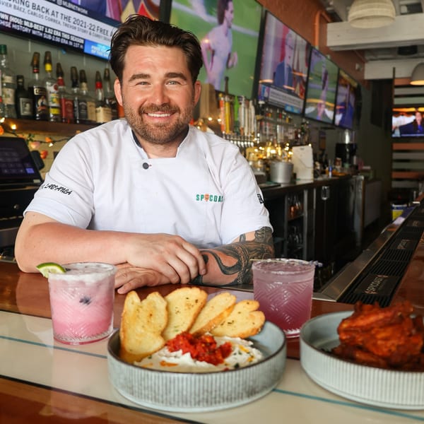 Chef in white coat smiling behind a bar with cocktails, dip, and wings at Spycoast Kitchen & Cocktails.