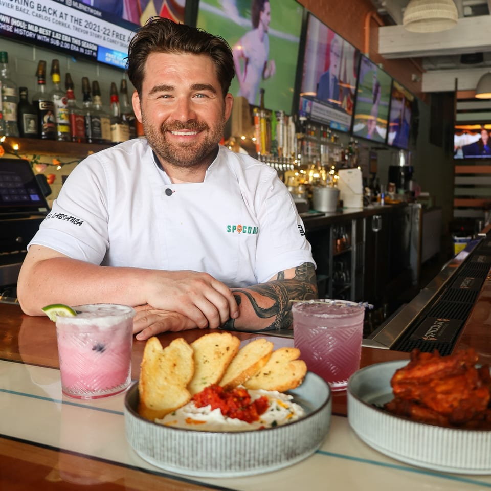 Chef in white coat smiling behind a bar with cocktails, dip, and wings at Spycoast Kitchen & Cocktails.
