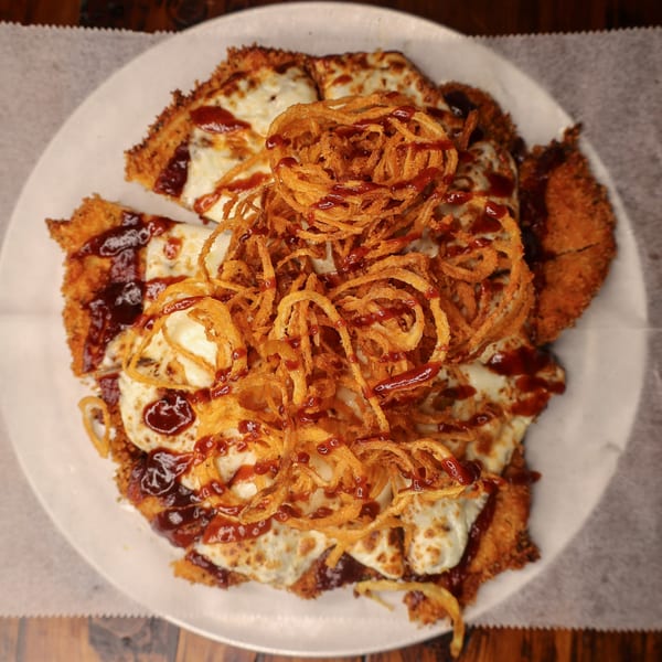 Crispy chicken schnitzel topped with melted mozzarella, BBQ sauce, and fried onion strings on a white plate.