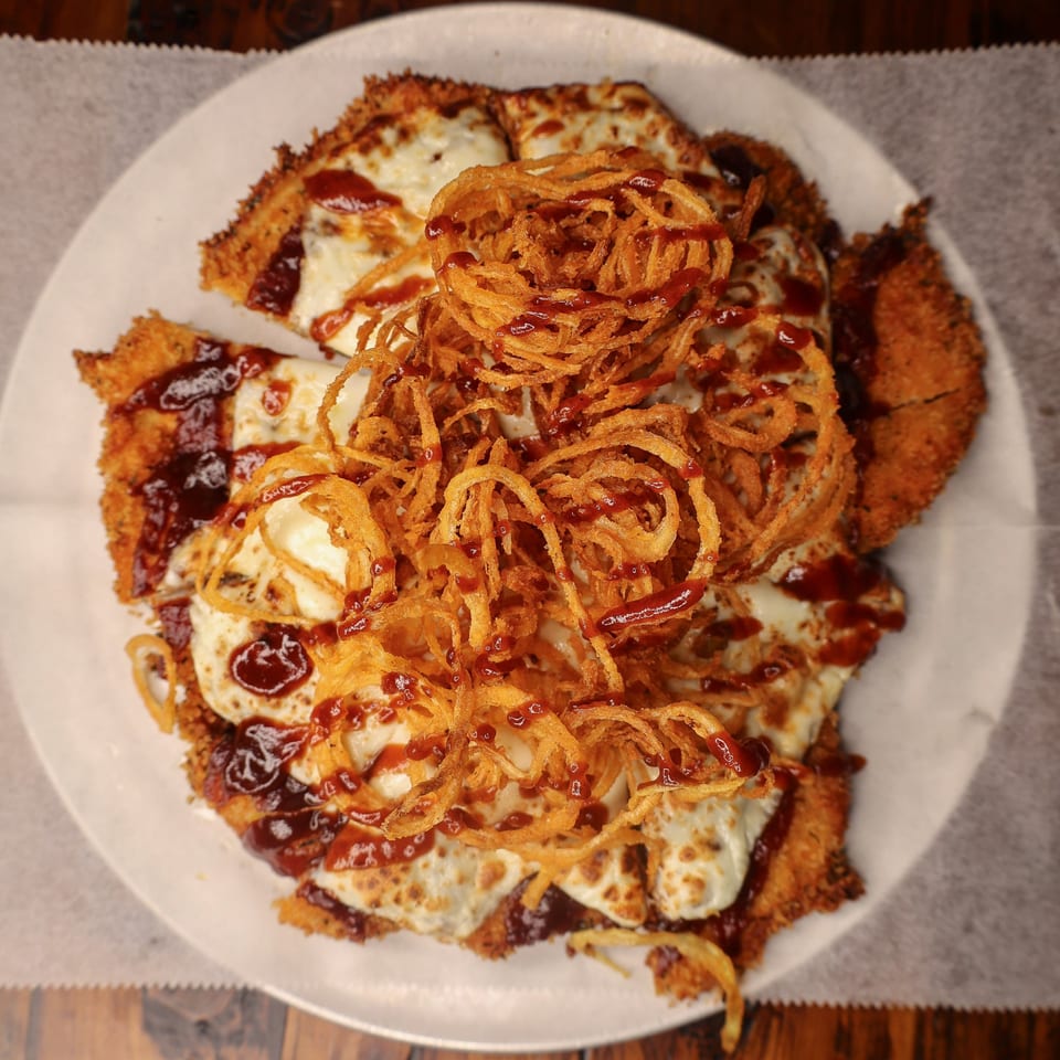 Crispy chicken schnitzel topped with melted mozzarella, BBQ sauce, and fried onion strings on a white plate.