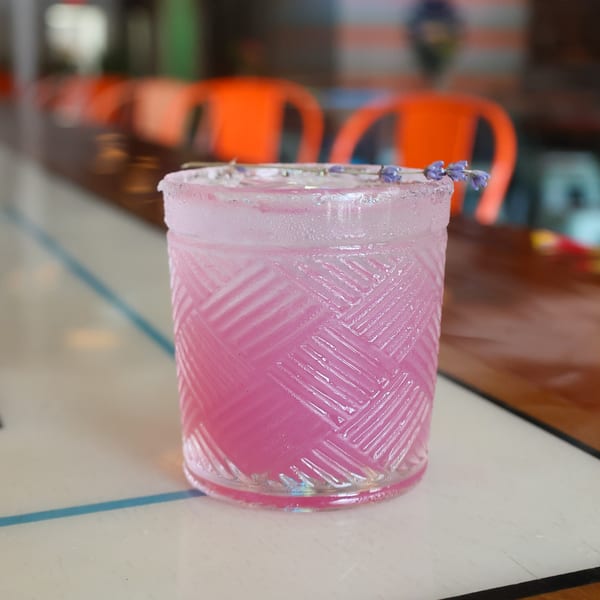 A pink cocktail in a textured glass, garnished with lavender, on a white bar top.